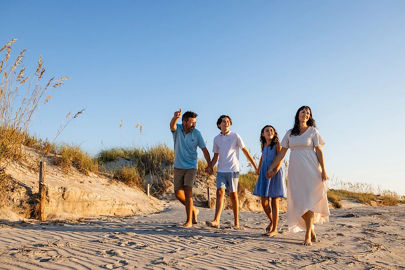 A family walks along a sunny beach, smiling and holding hands as they stroll toward the camera, carefree and joyful.