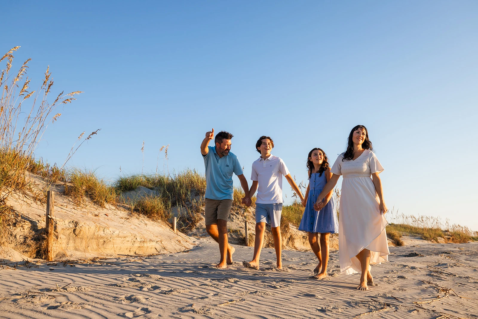 A family of four walks along a sandy beach at sunset, holding hands and smiling, enjoying a carefree stroll together.