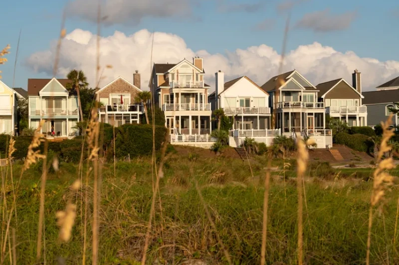 A row of colorful coastal houses on a windy bluff, sea grass in the foreground, blue sky with fluffy clouds behind.