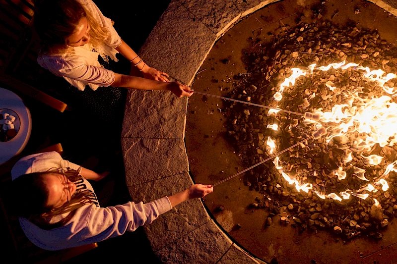 Two people at night, extending long skewers toward a blazing circular fire pit surrounded by rocks, gathering around a warm bonfire.