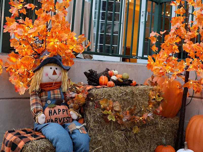 Autumn display: a scarecrow on hay bales with orange leaves, pumpkins, and seasonal decor at a porch.