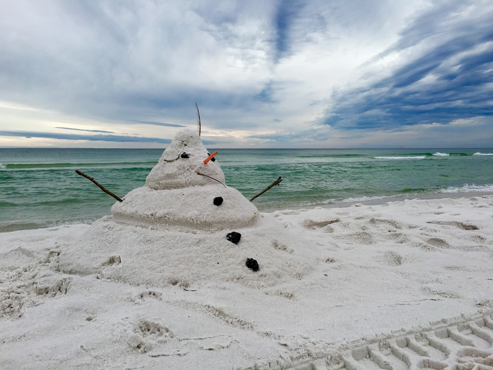 A tiny sand snowman on a sunny beach with a turquoise sea, two twig arms, a small carrot nose, and coal eyes, under a dramatic blue sky.