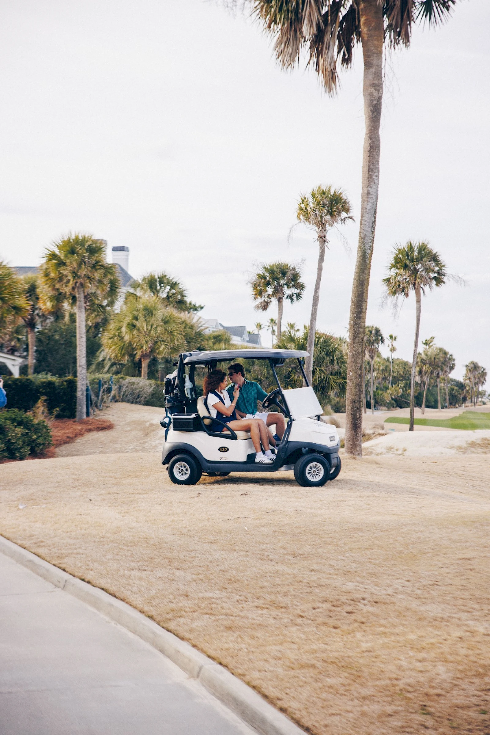 A small golf cart parked on a grassy path near palm trees; two people sit inside, casual vibe, sunny day, likely a resort or park.