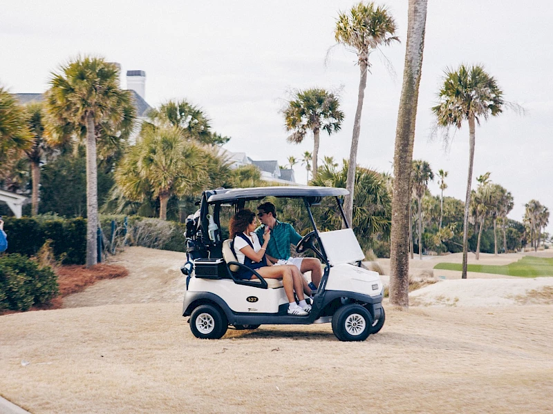 A small golf cart parked on a grassy path near palm trees; two people sit inside, casual vibe, sunny day, likely a resort or park.
