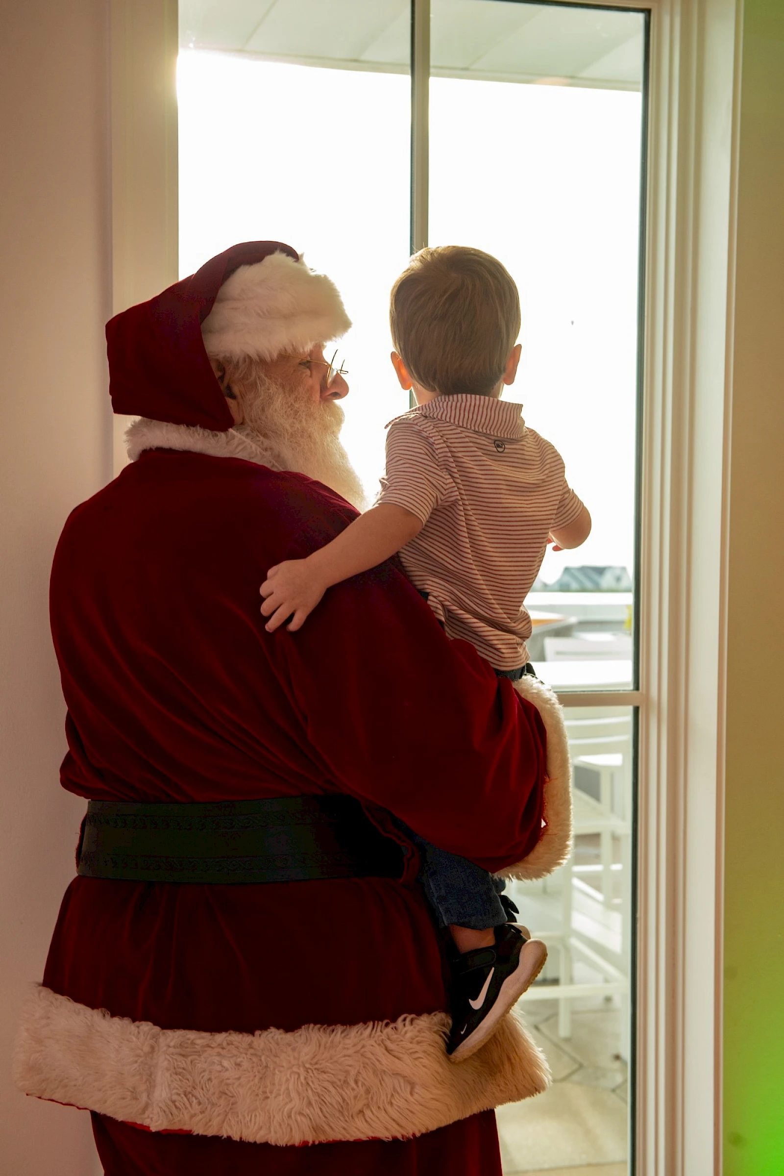 Santa Claus carrying a toddler by the window, sharing a warm holiday moment with a snowy view outside.