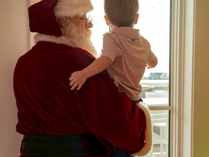 Santa Claus carrying a toddler by the window, sharing a warm holiday moment with a snowy view outside.