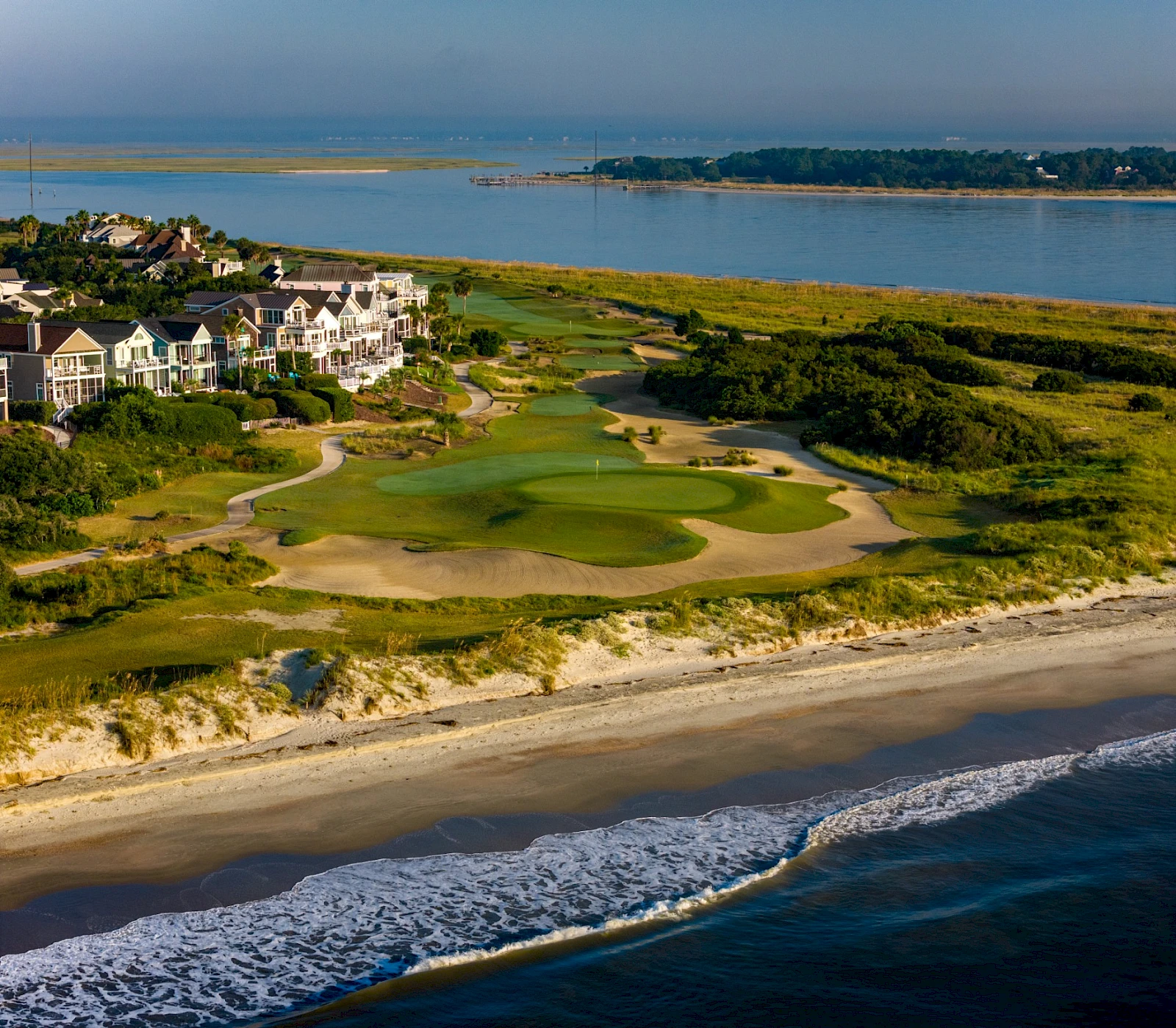 Coastal golf course with green fairways, sand traps, and water views; beachfront houses line the shore as waves wash onto the sandy beach.