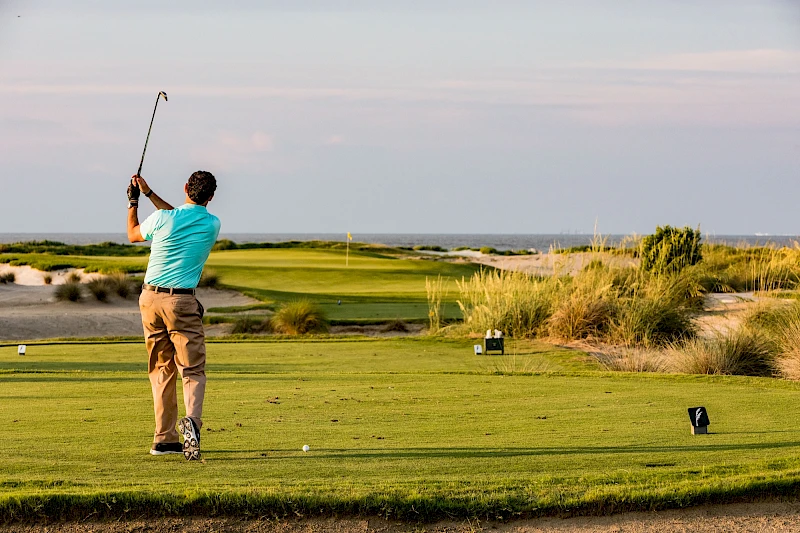 A golfer in a turquoise shirt and khaki pants swings a club on a green fairway, with dunes and shrubbery in the background.