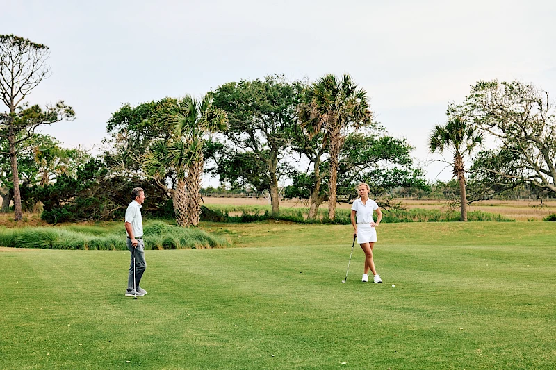 Two people on a golf course, one near the hole watching the other prepare to putt, with trees and a clear sky in the background.