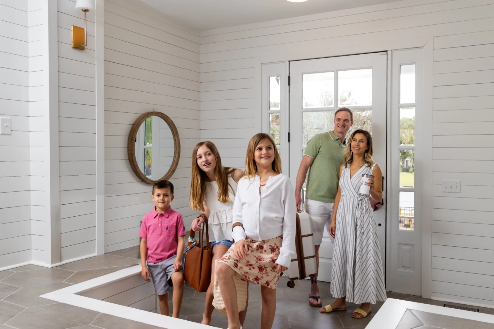 A family stands inside a modern entryway with white shiplap walls, glass doors, and a round mirror; kids in front, parents smiling behind them.
