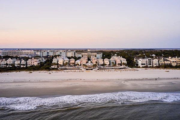A row of pastel beach houses lines a sandy shore, with calm ocean waves rolling in and a subtle sunset skyline above the horizon.
