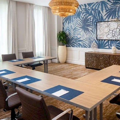 A modern conference room with a U-shaped wooden table, blue desk mats, brown chairs, a tropical blue wallpaper accent wall, and warm lighting.