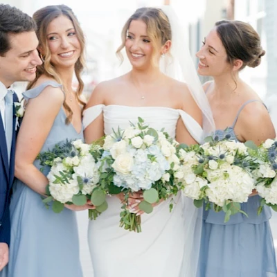 A wedding party photo with the bride in a white dress flanked by bridesmaids in light blue gowns, all smiling and holding bouquets.