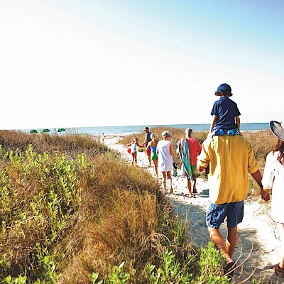 A group of people, including adults and kids, walk along a sandy path near the coast with tall grasses, heading toward the beach. End.