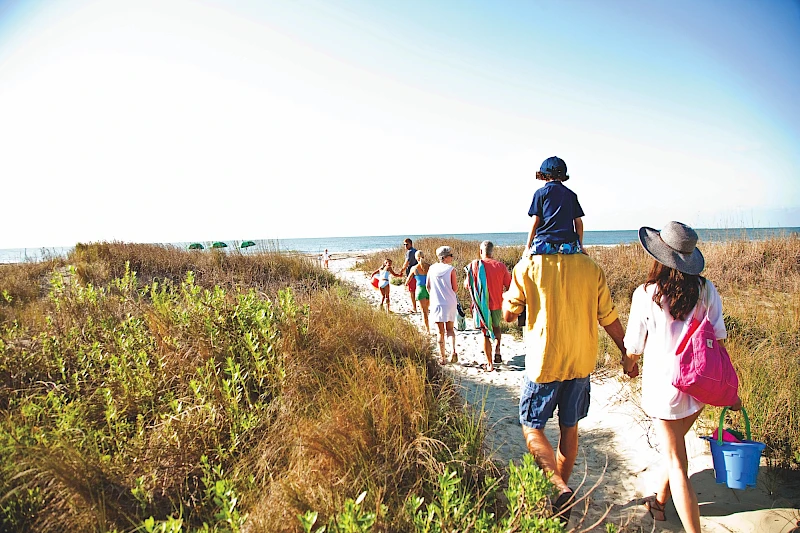 A group of people, including adults and kids, walk along a sandy path near the coast with tall grasses, heading toward the beach. End.