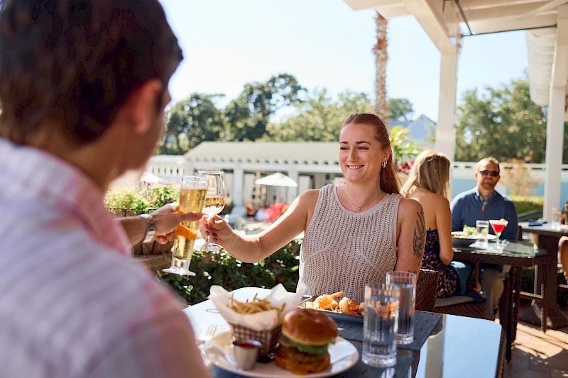 Two people clink glasses across a sunny outdoor restaurant table, with burgers, fries, and drinks in view, while smiling and enjoying a meal together.
