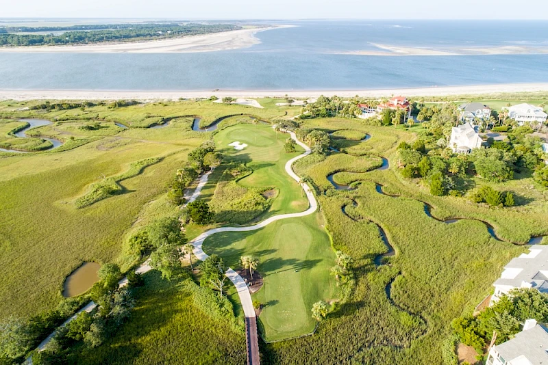 An aerial view of a coastal golf course with winding fairways, water hazards, and green greens, bordered by sandy beaches and marshy areas.