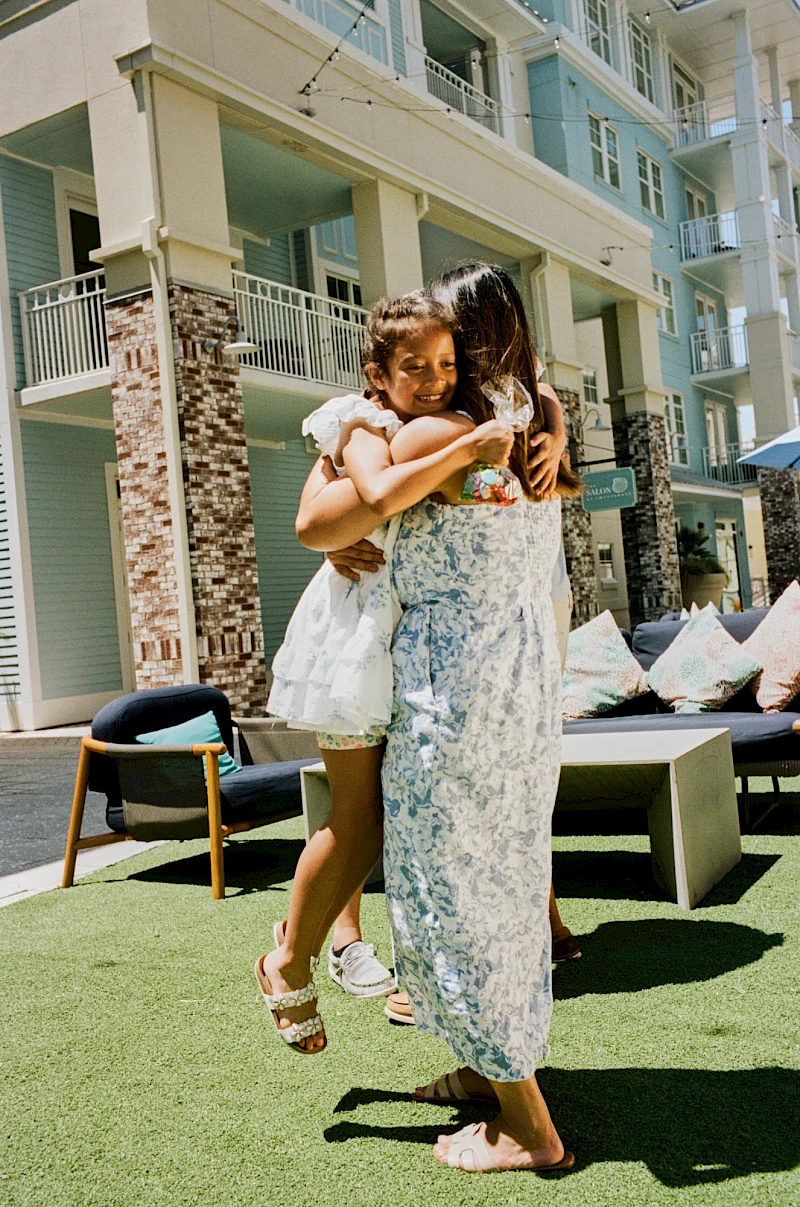 Two people share a warm hug on a sunny outdoor area near a modern hotel, with a child smiling while being lifted by an adult in a patterned dress.
