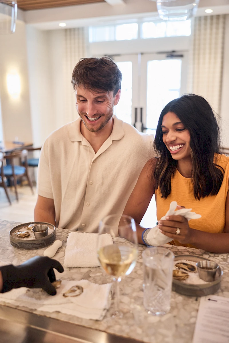 A smiling couple sits at a dining table with wine glasses and candles, enjoying a meal together in a bright, cozy restaurant.
