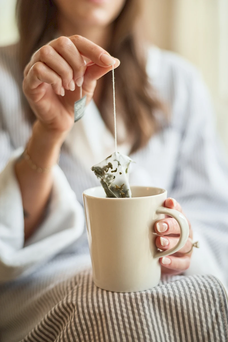 A person in a robe is stirring a mug with a tea bag or string, lifting it above a light-colored mug while sitting on a striped blanket.