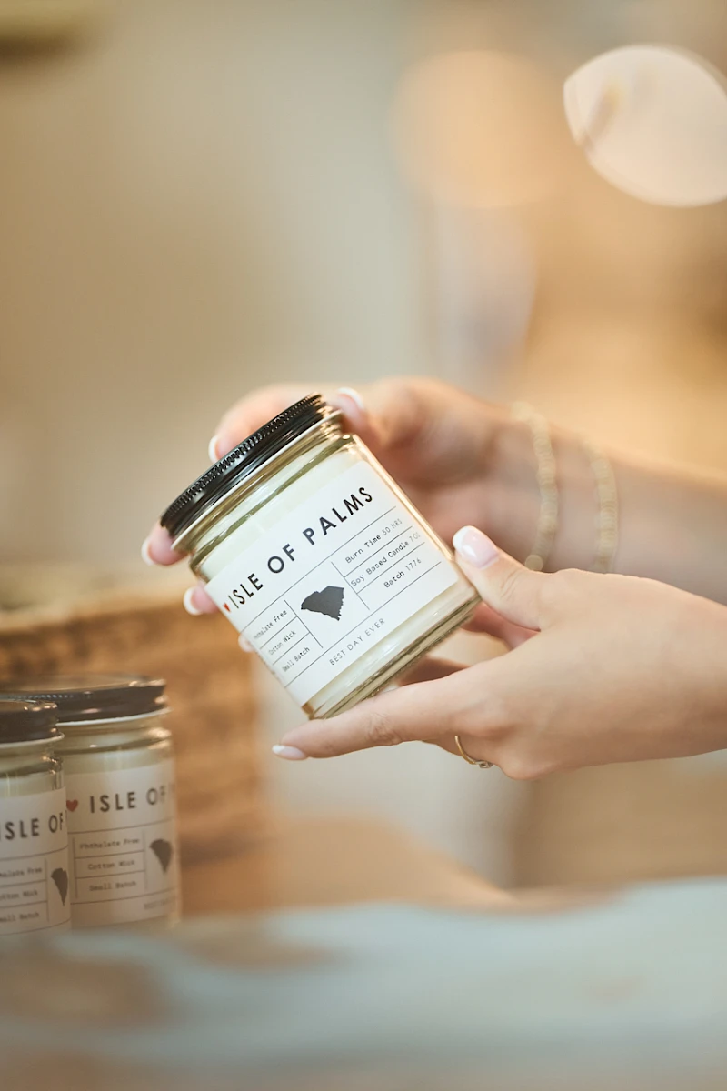 A person holding a jar labeled “House of Palms” in a cozy shop, with more jars on a blurred counter in the background.