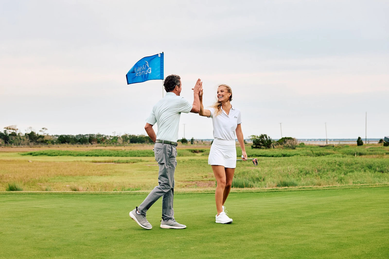 Two golfers on a greens, raising hands in a high-five as a blue flag waves in the background near a grassy field.