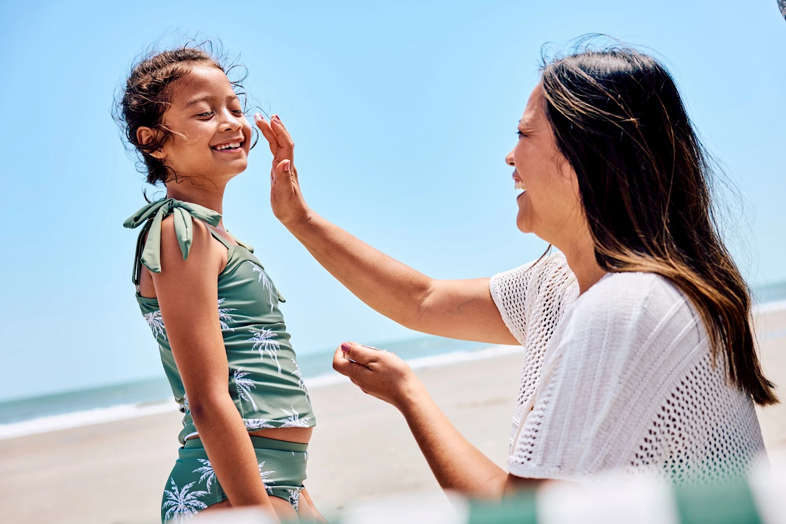 Two people on a sunny beach; an adult woman touches a smiling young girl’s face, sharing a playful moment by the shore.