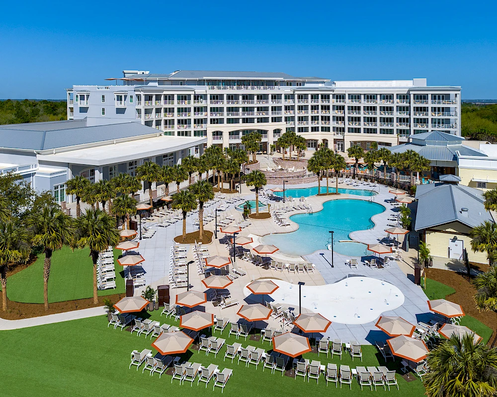 A resort with a large pool, lounge chairs, cabanas, palm trees, and a central building, set on sunny grounds.
