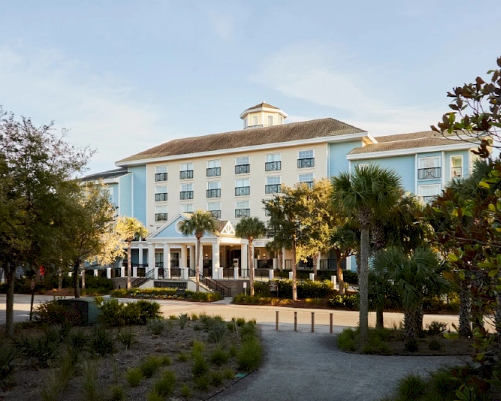 A light blue multi-story hotel or resort with a central entrance, palm trees, and a curved driveway, set among landscaped grounds.