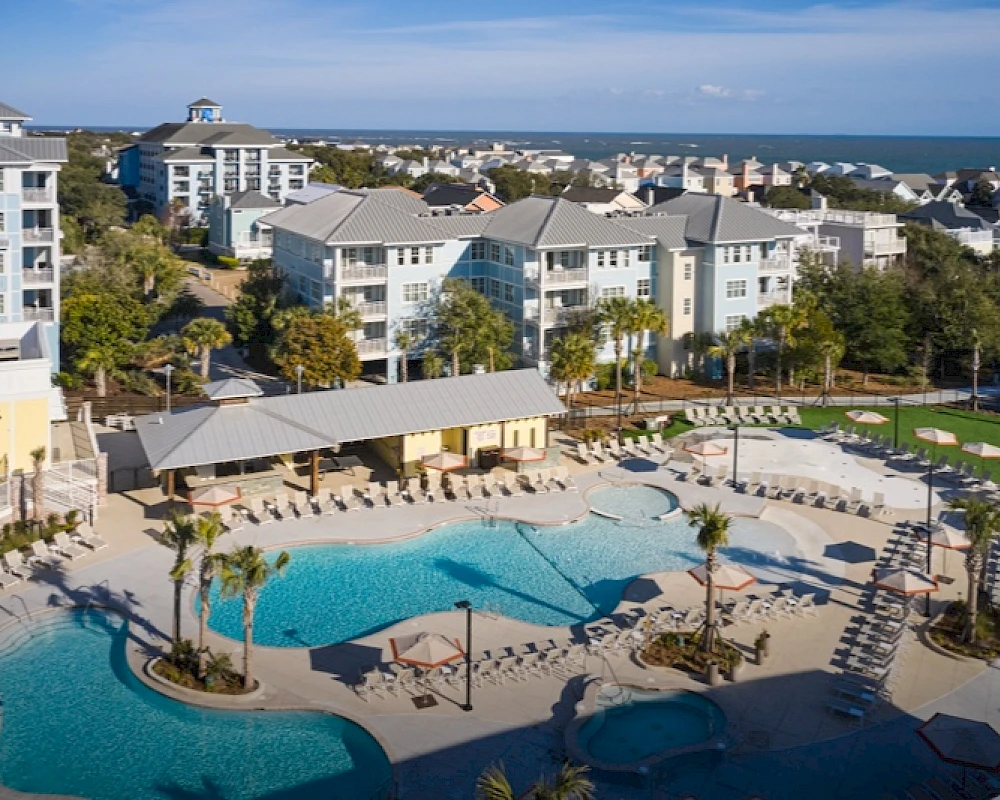 A coastal resort with a large outdoor pool, palm trees, lounge chairs, and multi-story white buildings; ocean backdrop and clear skies.