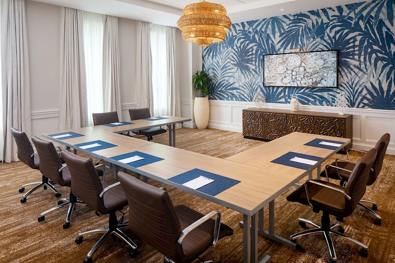 A modern conference room with a U-shaped table, blue place mats, white napkins, brown chairs, a bold blue botanical wallpaper, a large hanging light, and a decorative cabinet along the wall.