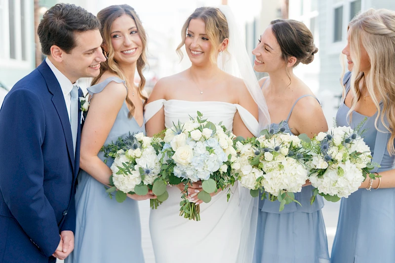 A wedding party: bride in white with bridesmaids in light blue dresses, holding white bouquet arrangements, smiling together.