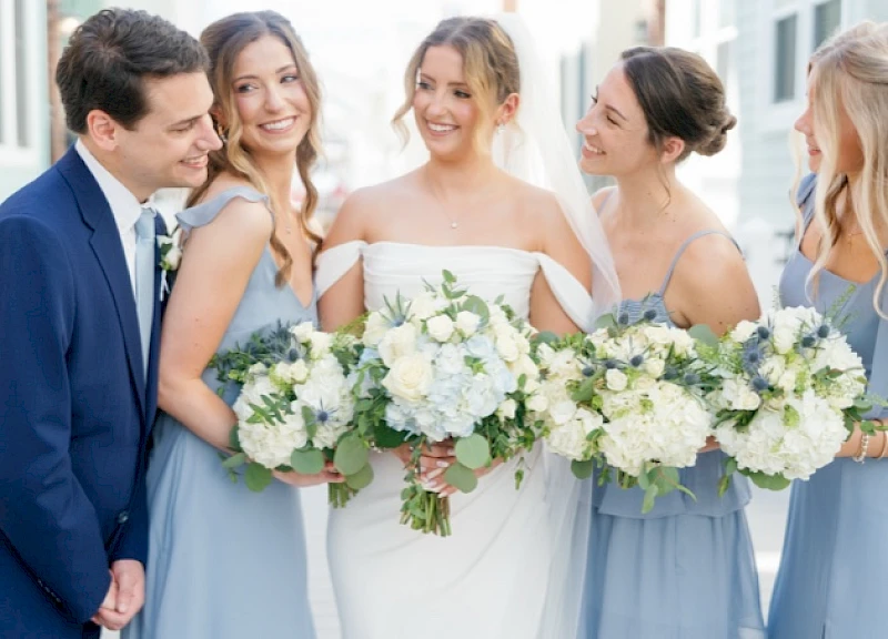 A wedding party: bride in white with bridesmaids in light blue dresses, holding white bouquet arrangements, smiling together.