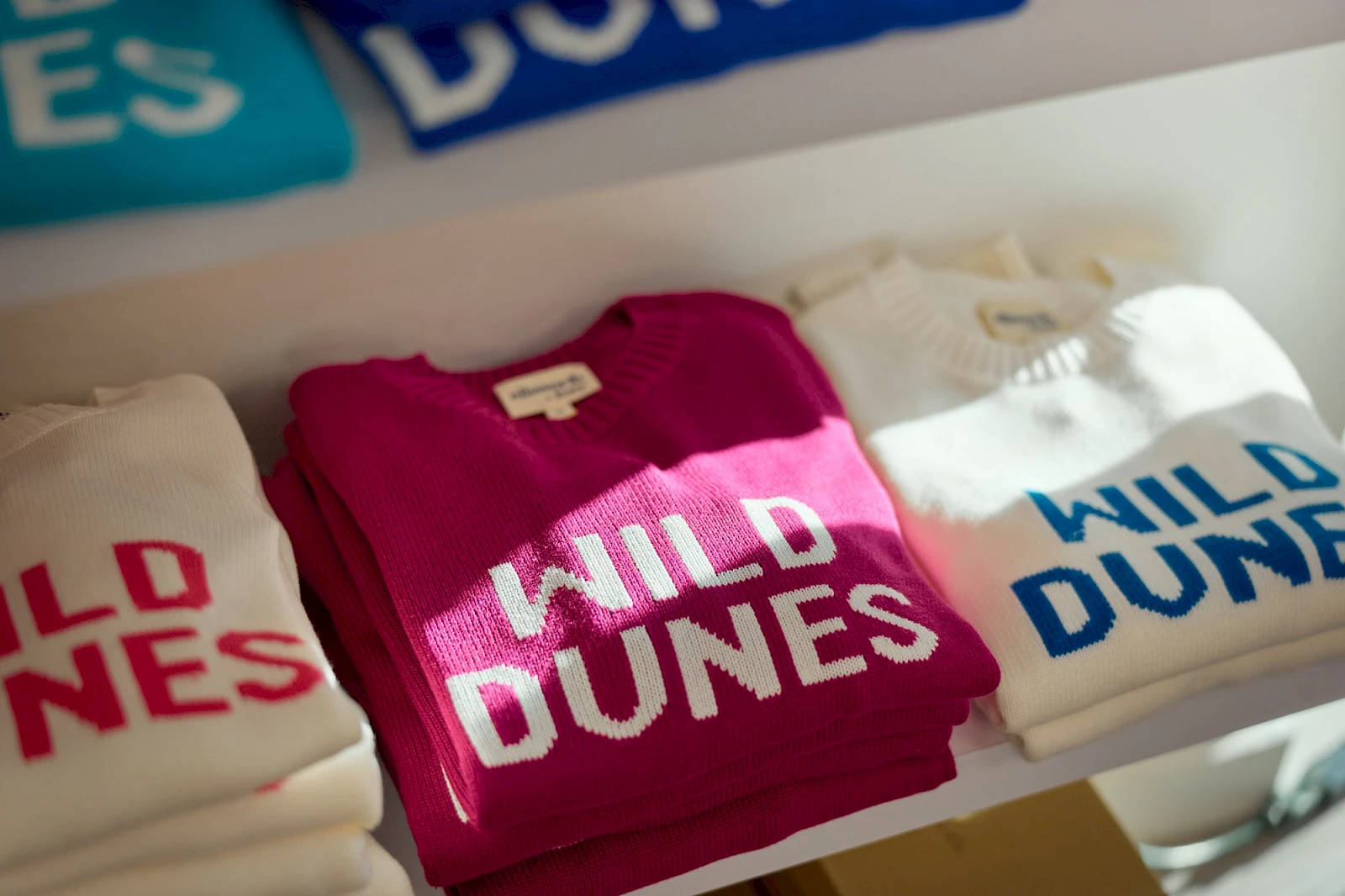 A row of folded sweaters with bold “WILD DUNES” lettering in white on cream, magenta, and white shirts, on a store shelf.