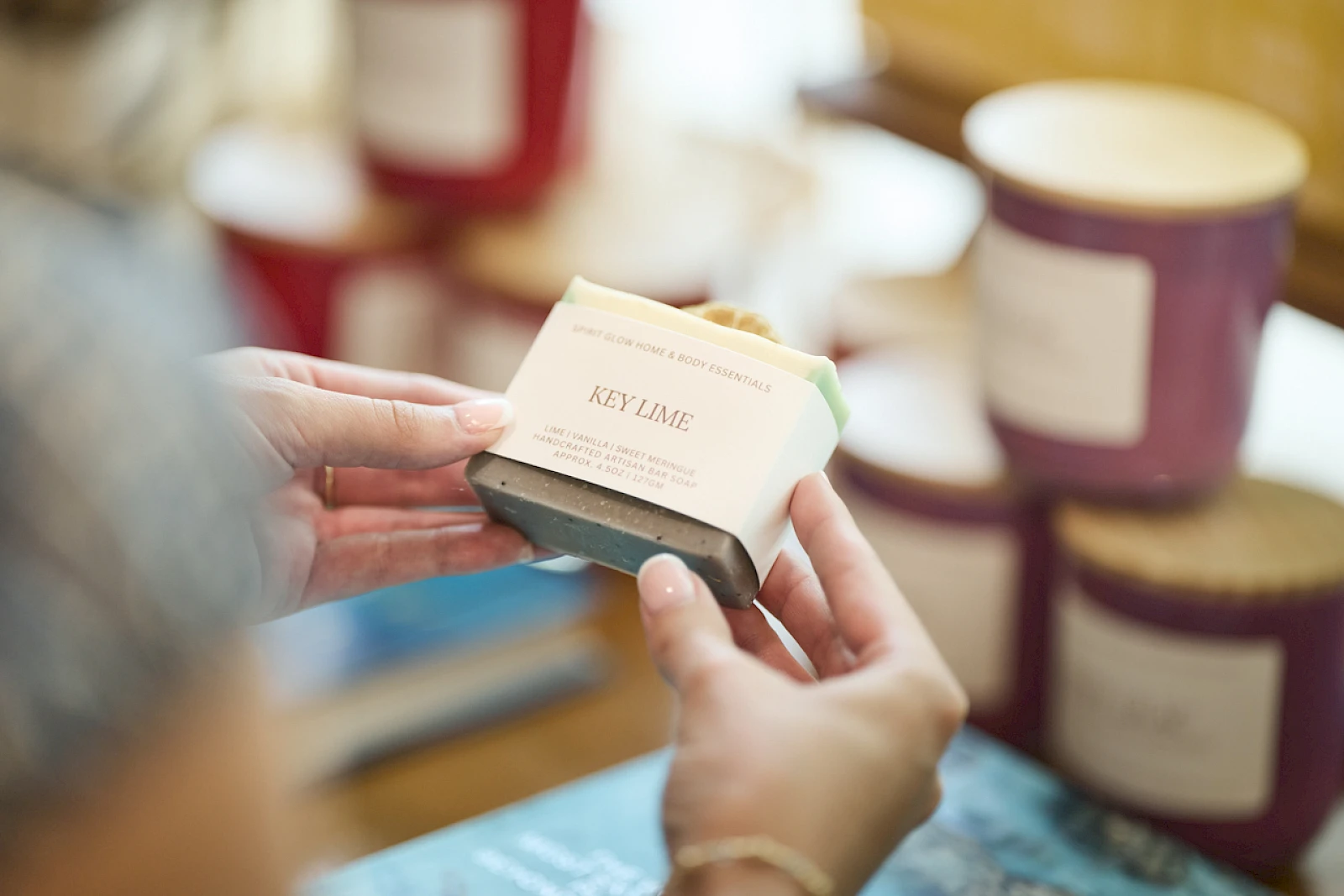 A person holds a small soap box labeled “Key Lime” in a cozy shop with jars and candles in the background.