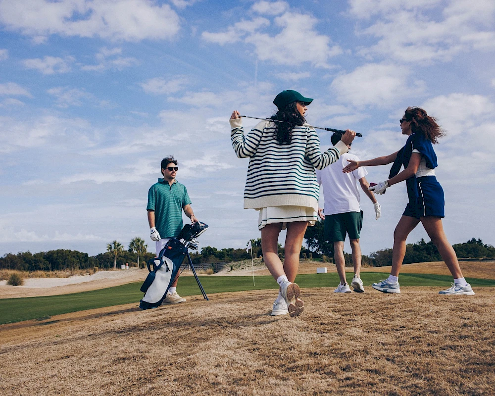 Friends on a sunny golf course: two people celebrate, others cheer with a bag and clubs nearby, blue sky, casual summer vibe.