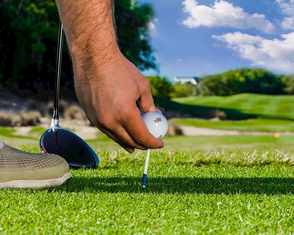 A close-up of a golfer placing a tee with a ball on the tee, foot in a cleat visible, teeing up on a sunny green fairway.