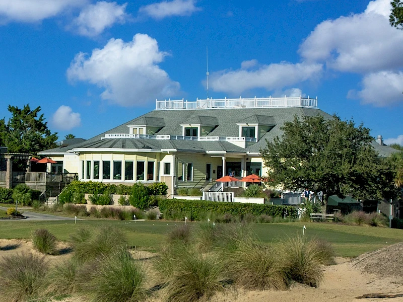 A large coastal-style house with a green lawn, patio seating, and shrubs under a bright blue sky with fluffy clouds.