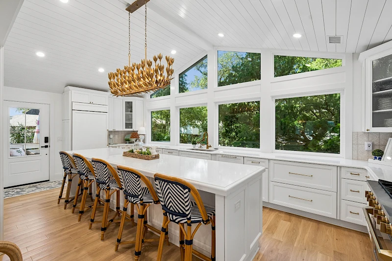 Modern white kitchen with a large island, pendant chandelier, bar stools, ample natural light, and a view of lush greenery outside.