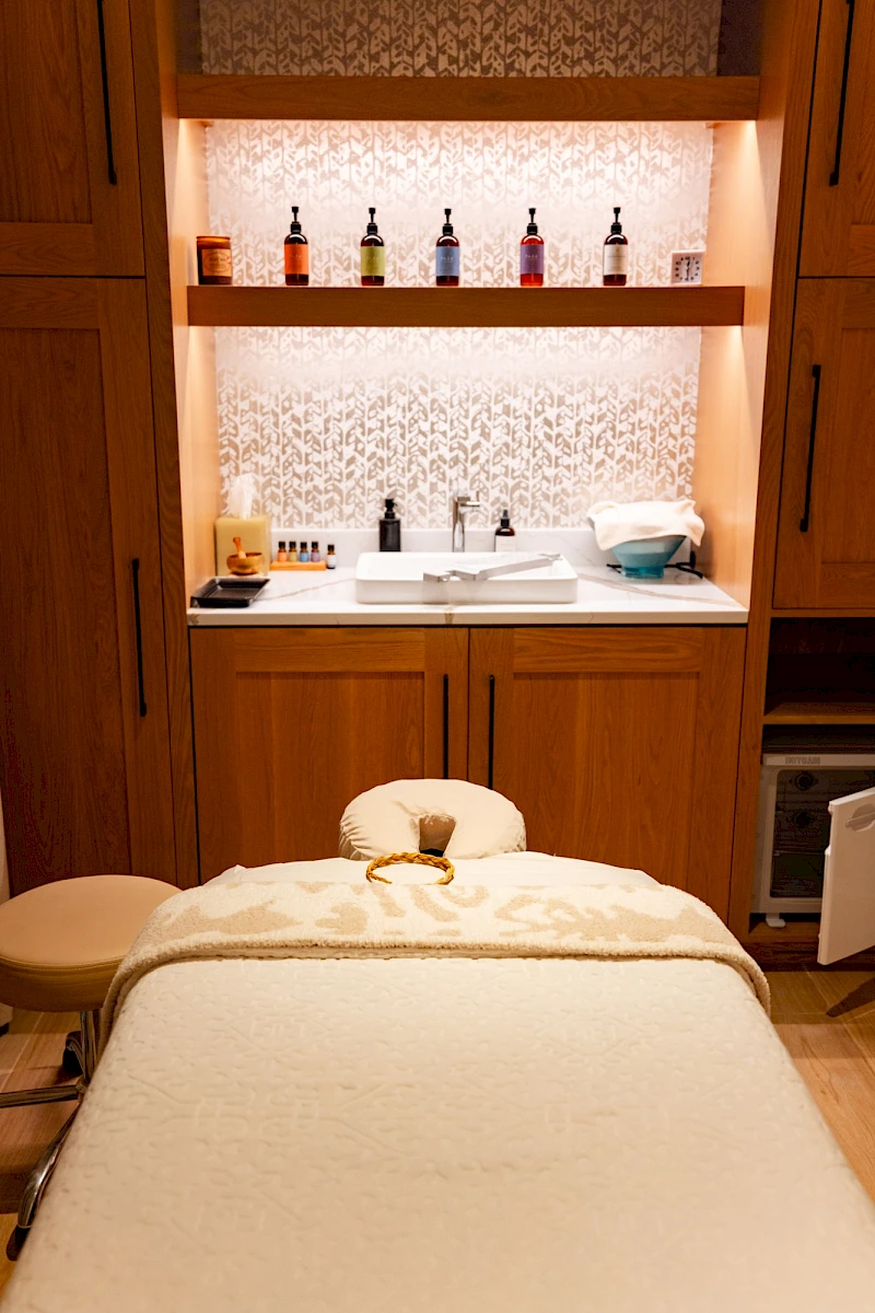 A spa treatment room with a massage bed, cabinets, and a sink area; shelves hold bottles and towels, a relaxing, warm-lit setup.