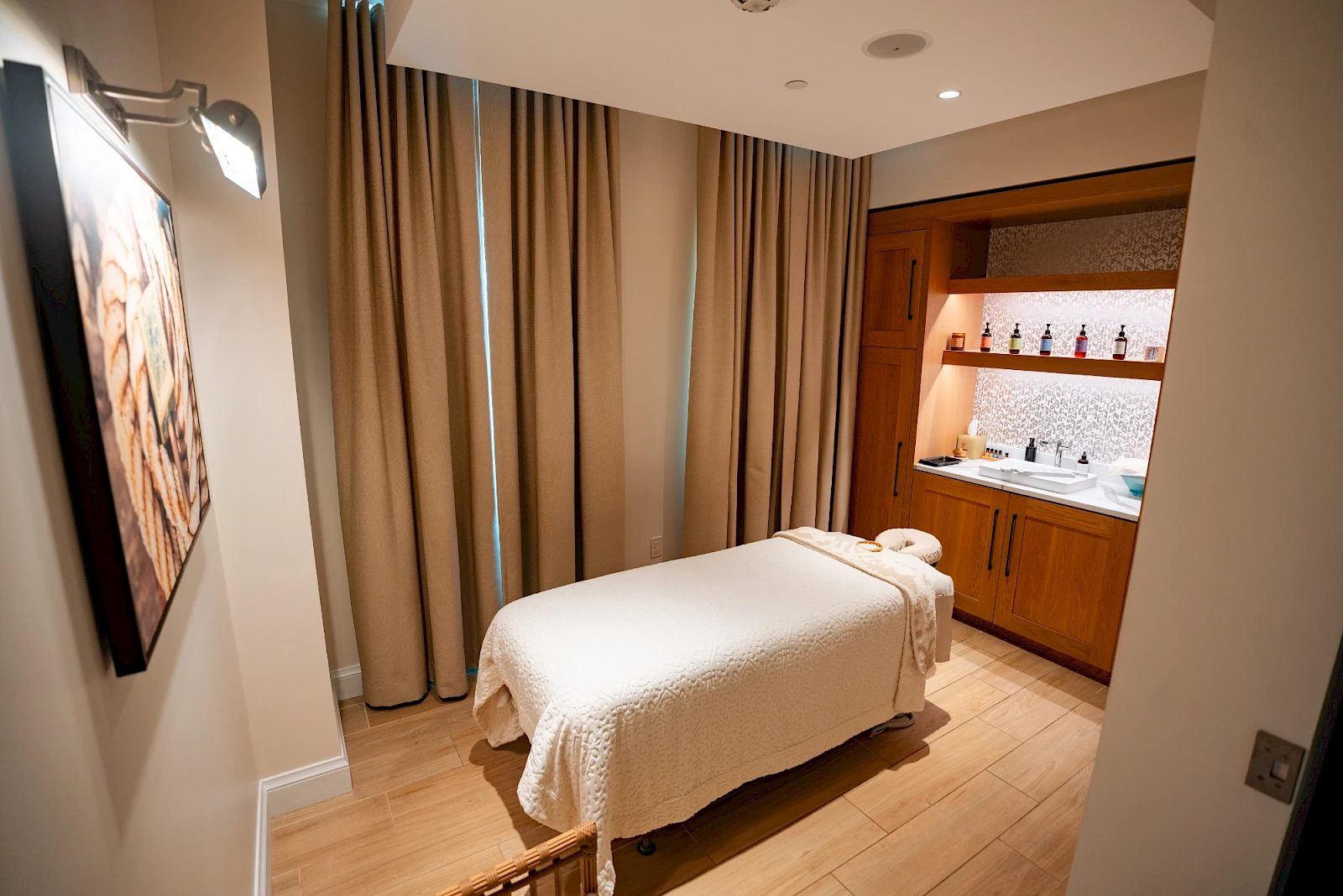 A small spa-style room with a single bed, beige blanket, wooden floors, curtains, and a compact kitchenette along the back wall. end.