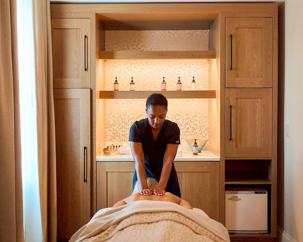 A massage therapist works on a client lying face down on a table in a tidy spa room with wooden cabinets and bottles on shelves.