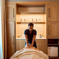 A massage therapist works on a client lying face down on a table in a tidy spa room with wooden cabinets and bottles on shelves.