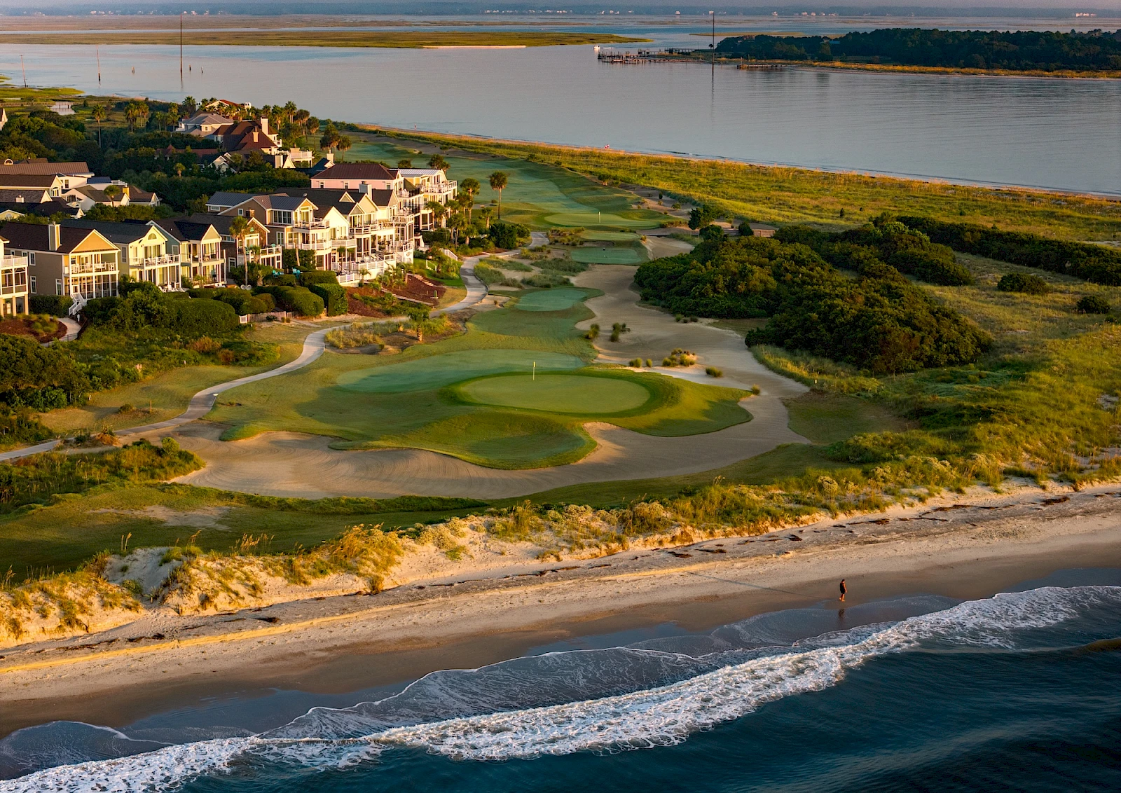 Coastal resort with a sandy beach, waterway, and a green golf course nestled among houses along the shore, waves washing onto the sand.