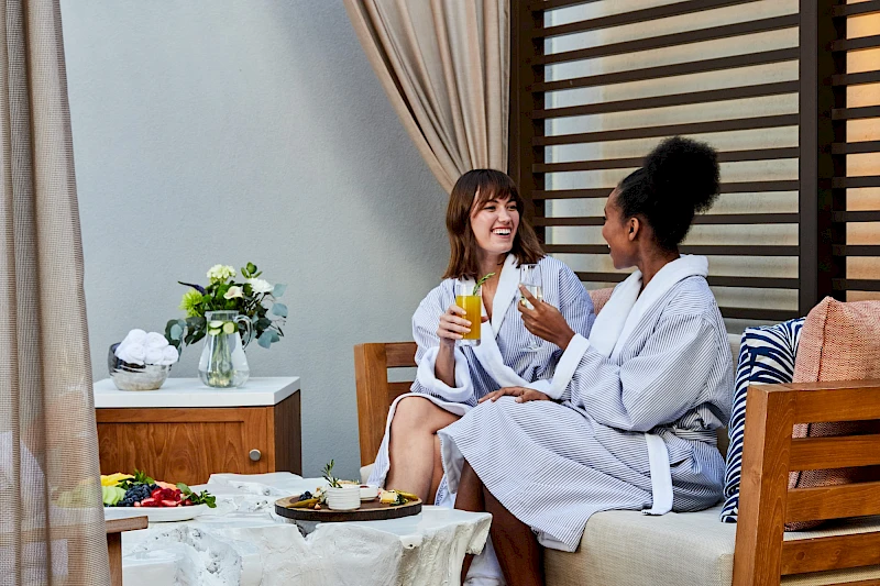 Two friends in robes toast with drinks on a balcony, enjoying snacks and a relaxed morning together.
