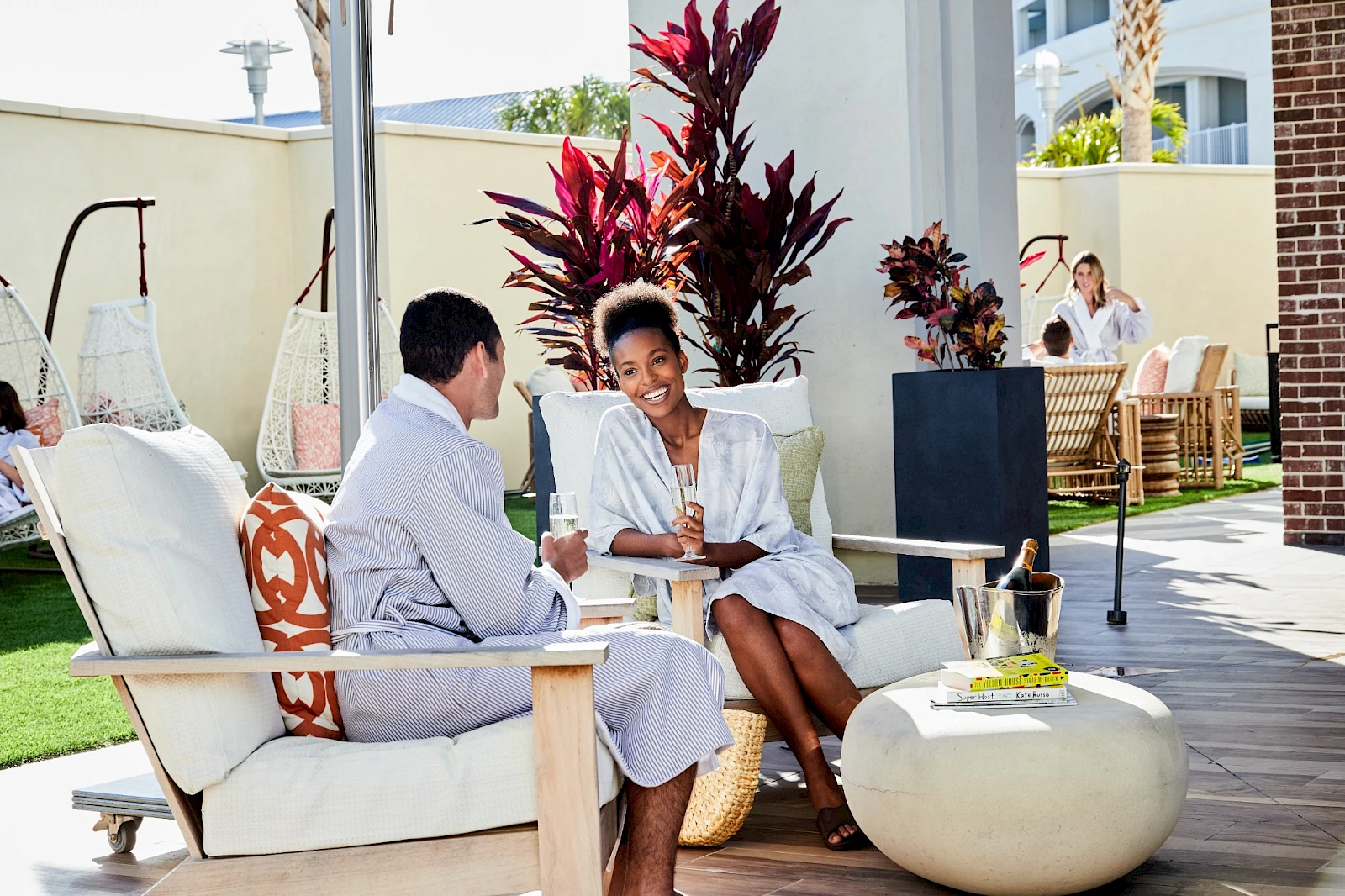 Two people relaxing on a patio, chatting and sipping drinks in white robes, with tropical plants and lounge seating.