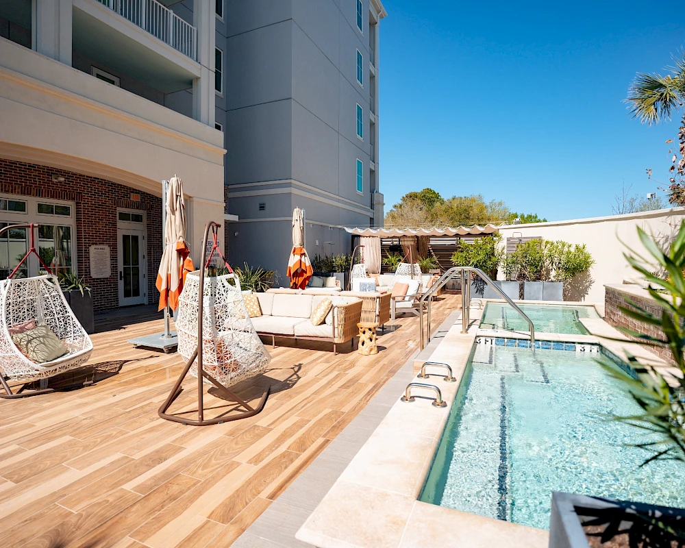 A sunny hotel pool deck with wooden flooring, hanging egg chairs, loungers, umbrellas, and a rectangular pool beside a tall building.