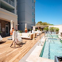 A sunny hotel pool deck with wooden flooring, hanging egg chairs, loungers, umbrellas, and a rectangular pool beside a tall building.