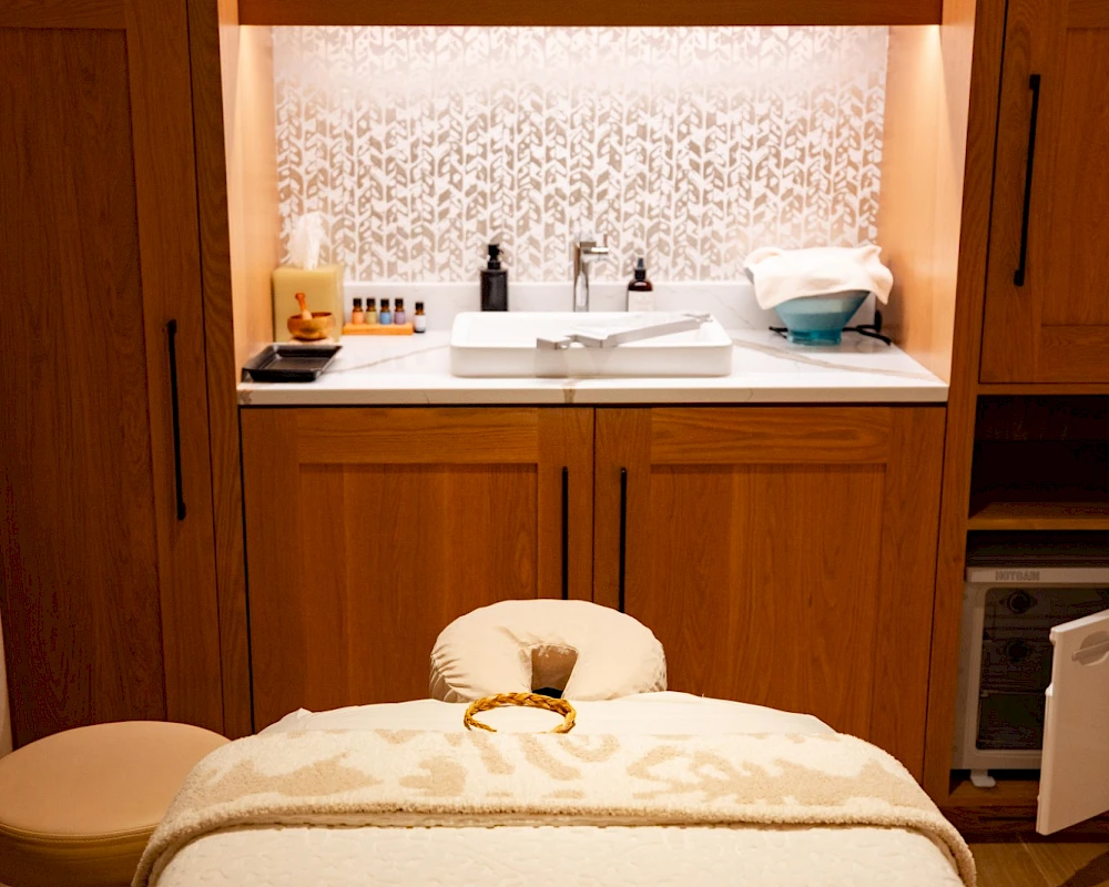 A cozy spa treatment room with a massage table, soft lighting, and a sink area behind wooden cabinets; bottles and supplies line the shelves. End.