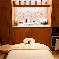 A cozy spa treatment room with a massage table, soft lighting, and a sink area behind wooden cabinets; bottles and supplies line the shelves. End.